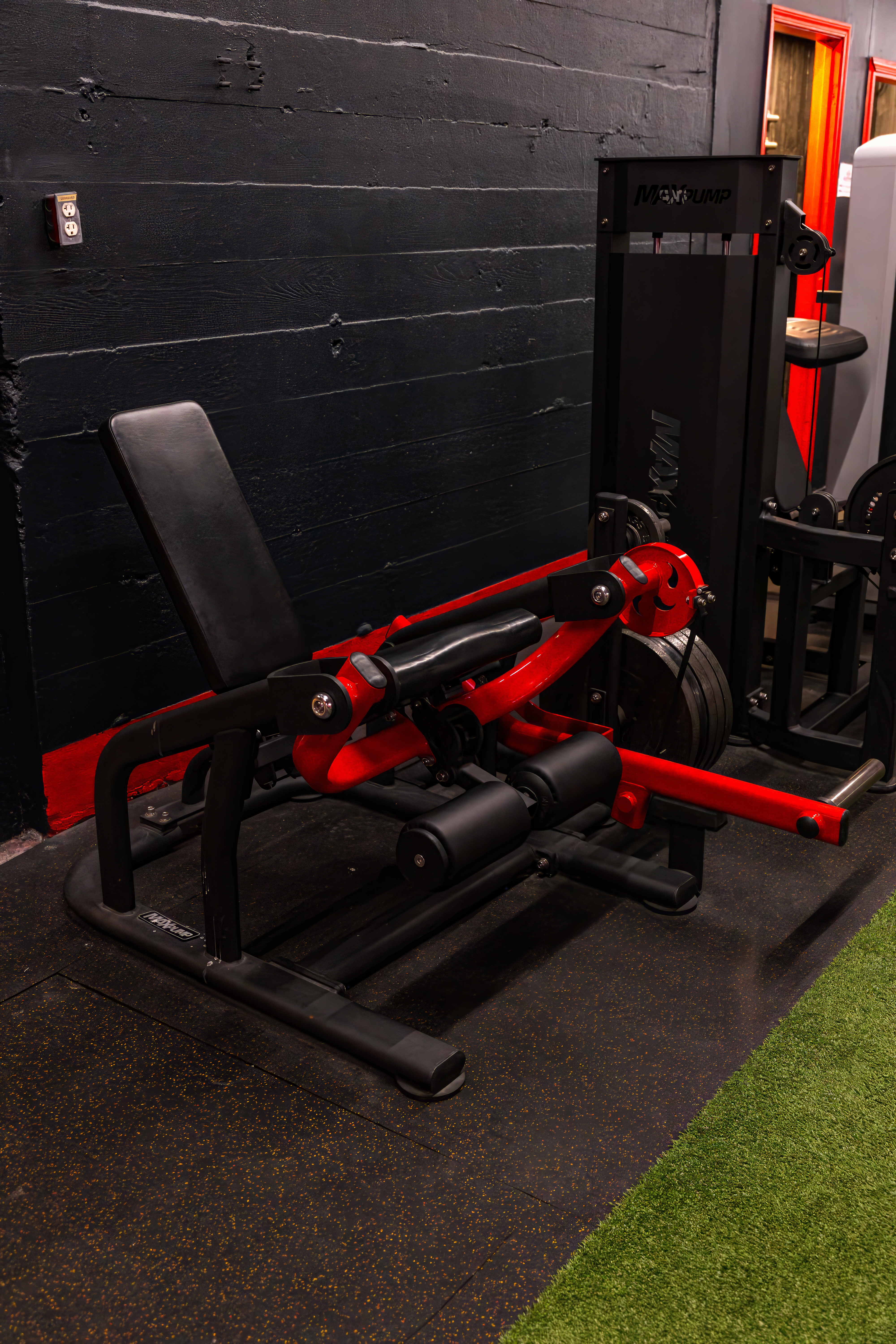 Seated leg curl machine with a distinctive red frame and black padding, situated on the border between the rubber flooring and the indoor turf